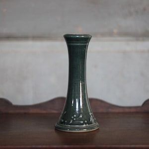 May include: A dark teal ceramic vase with a flared base and a slender, elongated body. The vase has a slightly textured surface and a decorative pattern near the base. The vase is displayed on a dark wooden surface.