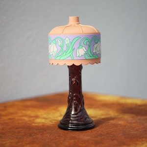 May include: A decorative table lamp with a floral patterned lampshade. The lampshade is pink, purple and green with white flowers. The base of the lamp is dark brown and has a textured design.