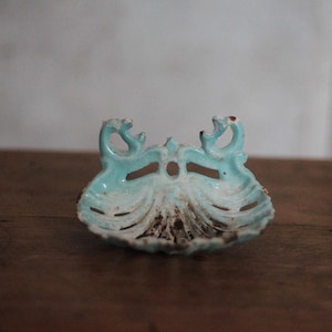 Può includere: Un piccolo piatto in ceramica turchese ornato con un motivo a conchiglia. Presenta due elementi decorativi curvi e un'apertura centrale. Il piatto è posto su una superficie di legno. Misura circa 15 cm di lunghezza.