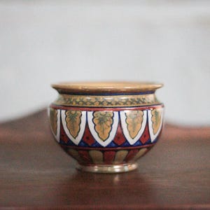Op de afbeelding: Een kleine, ronde keramische pot met een brede opening. De pot heeft een decoratief patroon van gouden, witte, rode en blauwe geometrische vormen. De rand en de basis zijn lichtbruin.