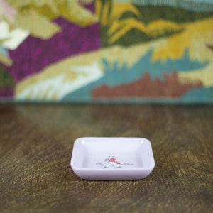 May include: A small, square, light pink ceramic dish with a white design of a person in a red jacket and black pants. The dish is sitting on a wooden surface.