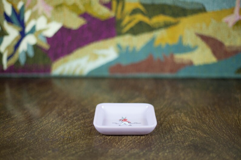 May include: A small, square, light pink ceramic dish with a white design of a person in a red jacket and black pants. The dish is sitting on a wooden surface.