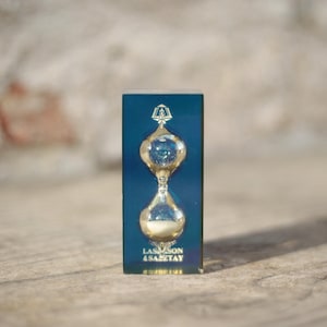 Könnte beinhalten: Ein rechteckiger blauer Acrylblock mit einer Sanduhr aus klarem Glas, die mit weißem Sand gefüllt ist. Die Oberseite der Sanduhr zeigt ein goldenes Emblem und der Text "LAS MASON & SANTAY" ist in Gold am unteren Rand des blauen Blocks aufgedruckt.