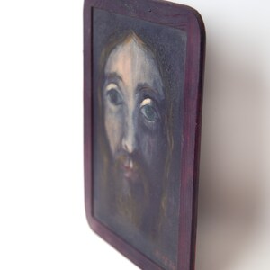 May include: A framed painting of a man's face with a beard. The painting is done in a dark, muted color palette with a focus on the man's eyes. The frame is dark wood with a slight purple hue.