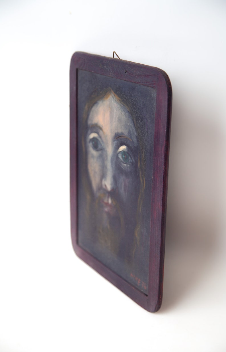 May include: A framed painting of a man's face with a beard. The painting is done in a dark, muted color palette with a focus on the man's eyes. The frame is dark wood with a slight purple hue.
