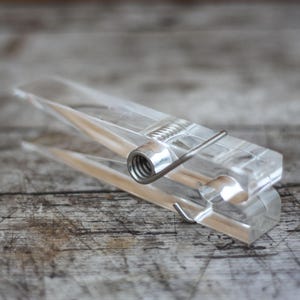 Könnte beinhalten: Eine transparente Wäscheklammer aus Kunststoff mit einer silbernen Metallfeder und einem Scharnier. Die Klammer ist klar und gibt den Blick auf die Holzoberfläche frei. Das Design ist einfach und funktional, zum Aufhängen von Wäsche oder anderen Gegenständen.