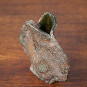 K&ouml;nnte beinhalten: Eine verwitterte, abstrakte Skulptur mit einer strukturierten, bronzefarbenen Au&szlig;enseite und Flecken von gr&uuml;nem Gr&uuml;nspan. Die Skulptur hat eine organische, asymmetrische Form, die m&ouml;glicherweise einem stilisierten Blatt oder Bl&uuml;tenblatt &auml;hnelt. Das Innere zeigt einen dunkleren Gr&uuml;nton.
