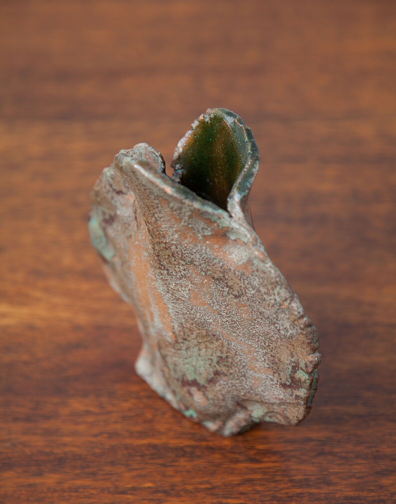K&ouml;nnte beinhalten: Eine verwitterte, abstrakte Skulptur mit einer strukturierten, bronzefarbenen Au&szlig;enseite und Flecken von gr&uuml;nem Gr&uuml;nspan. Die Skulptur hat eine organische, asymmetrische Form, die m&ouml;glicherweise einem stilisierten Blatt oder Bl&uuml;tenblatt &auml;hnelt. Das Innere zeigt einen dunkleren Gr&uuml;nton.