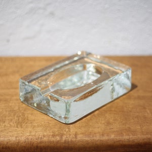 Op de afbeelding: Een rechthoekige asbak van helder glas. De asbak staat op een houten oppervlak.