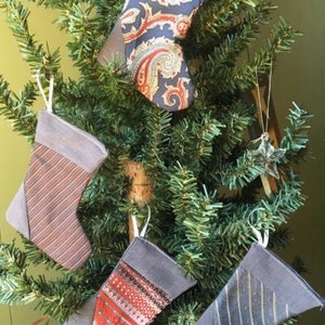 May include: Four small Christmas stockings with different patterns hanging on a green Christmas tree. The stockings are made of fabric and have a gray, red, and white color scheme.