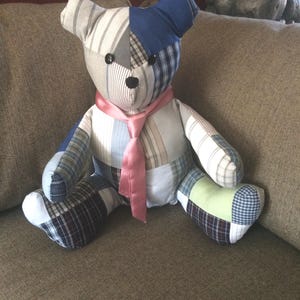 May include: A patchwork teddy bear with a pink satin bow tie. The bear is made from different fabrics, including plaid, striped, and solid colors. The bear is sitting on a brown couch.