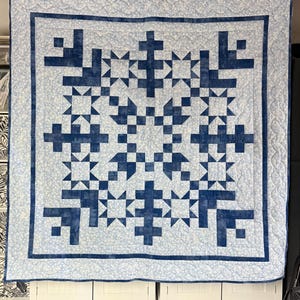 May include: A square quilt featuring a geometric pattern of dark blue fabric blocks arranged in star and cross shapes against a light blue and white floral patterned background, finished with a dark blue border and intricate quilted stitching.