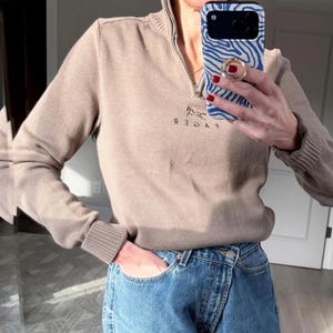 May include: A tan-colored sweater with a quarter-zip neckline and ribbed cuffs. The sweater features a small embroidered horse logo and text. The person in the image is wearing blue jeans.