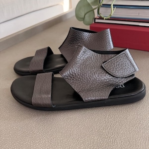 May include: A pair of metallic charcoal gray sandals featuring a textured leather upper, a single front strap, and a wide ankle strap with a hook-and-loop closure. The sandals have a smooth black footbed and a flat black sole for a simple design.