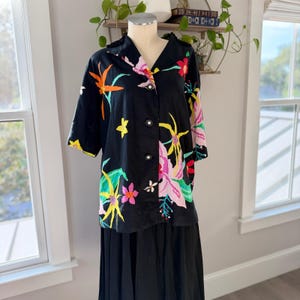 May include: A black short-sleeved button-down shirt featuring a vibrant floral print with yellow, orange, pink, and green flowers. The shirt has a notched collar and three large black buttons down the front. It is displayed on a mannequin.