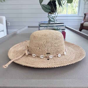 May include: A beige woven sun hat with a wide brim. The hat is adorned with a string of white seashells around the crown and has a long tie. The hat is designed for sun protection and fashion.