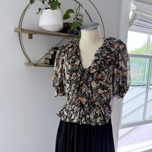 May include: A floral print blouse with a V-neck and short sleeves. The blouse features a ruffled neckline and a gathered waist. The bottom half of the garment is a solid black color. The blouse is displayed on a mannequin.