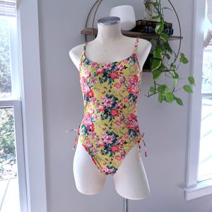 May include: A one-piece swimsuit featuring a vibrant yellow base color with a dense floral pattern in shades of pink, red, and green. The garment includes thin shoulder straps, a scoop neckline, and adjustable side ties at the hip area.
