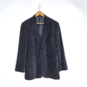 Op de afbeelding: Een donkergrijze blazer met een sluiting met twee knopen, een revers met inkeping en twee voorzakken. De blazer hangt aan een houten kleerhanger tegen een witte achtergrond. De stof lijkt zacht en getextureerd.