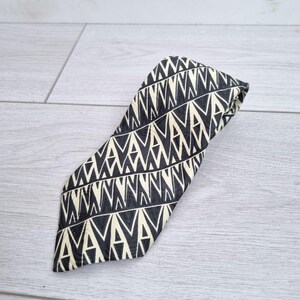 Puede incluir: Una corbata doblada con un patrón geométrico repetitivo en negro y crema. El patrón consiste en una serie de triángulos conectados y formas similares a letras. La corbata está hecha de una tela suave.