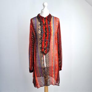 May include: A long-sleeved, sheer tunic with a vibrant, patterned design. The tunic features vertical stripes in red, orange, purple, and black, with a mandarin collar and decorative buttons. The garment is displayed on a white mannequin.