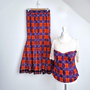 May include: A vintage two-piece outfit featuring a skirt and a top. The skirt is a long, pleated style with a plaid pattern in orange, purple, and white. The top has a white lace bodice and matching plaid pattern. The outfit is displayed on a white background.