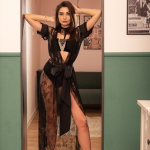 May include: A woman wearing a black lace robe with a satin sash and matching lingerie. She is standing in a doorway with a green wall on one side and a pink wall on the other.