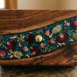You said
Describe this image in less word

Gemini said
A dark wood box with a wavy teal resin inlay containing dried purple roses, green leaves, and gold flakes. The lower corner features an engraved rose logo and the name "Jenkins."