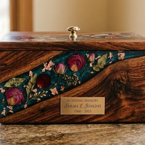 Puede incluir: Urna de madera con pomo de lat&oacute;n e incrustaciones de resina turquesa. La incrustaci&oacute;n presenta flores secas y hojuelas doradas. Una placa de lat&oacute;n dice "In Loving Memory Sarah L. Jenkins 1960-2023".