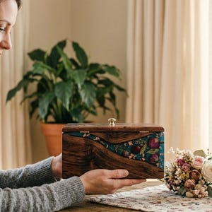 Puede incluir: Una mujer sostiene una caja de recuerdos de madera con una incrustaci&oacute;n de resina floral y turquesa. La caja tiene un cierre dorado. Un retrato enmarcado y un ramo de flores est&aacute;n sobre una mesa cercana.