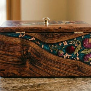 A handcrafted wooden keepsake box featuring a wavy teal resin inlay embedded with dried pink roses, botanical sprigs, and gold leaf flakes. The box has a polished natural grain, a hinged lid, and a decorative gold knob.