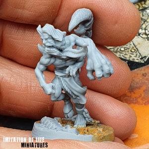 Könnte beinhalten: Graue Miniaturfigur einer gehörnten humanoiden Kreatur mit muskulösem Körperbau, Ketten an den Knöcheln und einem fließenden Gewand. Die Figur steht auf einem strukturierten Sockel mit dem Text "IMITATION OF LIFE MINIATURES".