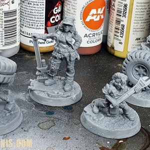 Może przedstawiać: Cztery niepomalowane figurki, w tym postać żeńska z kluczem, postać z głową w kształcie opony i postać żeńska trzymająca młotek. Figurki są na okrągłych podstawach, gotowe do malowania. W tle znajdują się słoiki z farbą.