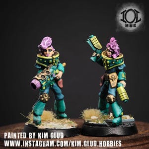 Op de afbeelding: Twee miniatuurbeeldjes van vrouwelijke personages in teal en gouden harnas, elk met een wapen. Ze hebben paars haar en staan op een kleine basis. De tekst "PAINTED BY KIM GLUD" en een websiteadres zijn zichtbaar.