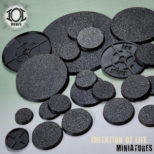 Puede incluir: Una colección de bases en miniatura negras y texturizadas de varios tamaños y formas. Algunas bases tienen diseños detallados, mientras que otras son lisas. Las bases están dispuestas sobre una superficie verde claro. El texto "IMITATION OF LIFE MINIATURES" es visible.