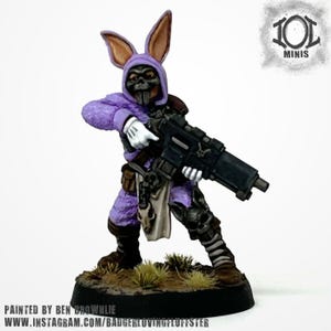 Può includere: Figurina in miniatura di un soldato in costume da coniglio viola, con un fucile nero. La figura indossa una maschera e guanti bianchi. La base è marrone con erba verde. Testo in basso: "PAINTED BY BEN BROWNLEE" e un indirizzo web.