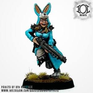 Könnte beinhalten: Miniaturfigur einer Person in einem blauen Hasenkostüm, die ein schwarzes Gewehr hält. Die Figur hat blondes Haar und ist detailliert bemalt. Der Sockel ist dunkel mit kleinen Grasbüscheln. Die Figur ist mit "101 MINIS" beschriftet.