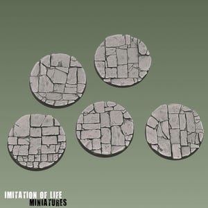 Pode incluir: Cinco bases circulares em miniatura com um padrão de calçada de pedra texturizada em tons de cinza. Cada base exibe um arranjo único de formas de pedra retangulares e irregulares. O texto "IMITATION OF LIFE MINIATURES" é visível.