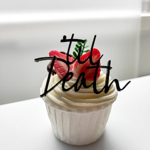 May include: A white cupcake with white frosting and a black acrylic topper that says "Til Death". The cupcake is decorated with a red strawberry.