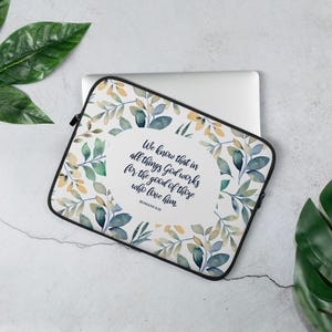 Puede incluir: Funda para portátil con un diseño floral de hojas verdes, azules y amarillas. Tiene una cremallera negra y un óvalo blanco en el centro con el texto "We know that in all things God works for the good of those who love him. Romans 8:28."