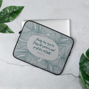 Puede incluir: Una funda gris para portátil con un estampado de hojas tropicales y el texto "fixing our eyes on Jesus, the pioneer and perfecter of faith. Hebrews 12:2." La funda tiene una cremallera y un ribete negros.