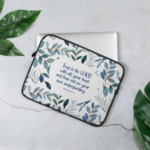 Könnte beinhalten: Eine Laptop-Hülle mit weißem Hintergrund, verziert mit einem Aquarelldesign aus blauen und grünen Blättern. Der Text "Trust in the LORD with all your heart and lean not on your own understanding" ist abgebildet. Sie hat einen schwarzen Reißverschluss.
