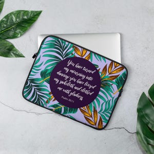 Puede incluir: Funda para portátil con un estampado de hojas tropicales en tonos verdes, amarillos y morados. Un diseño circular en el centro muestra una cita del Salmo 30:11 en texto blanco. La funda tiene una cremallera negra.