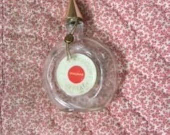 Puede incluir: Una botella de vidrio transparente y redonda con un tapón dorado y una etiqueta blanca y roja. La botella tiene una forma pequeña y circular y un cuello largo y delgado. Una etiqueta está adherida a la botella. La botella está vacía.