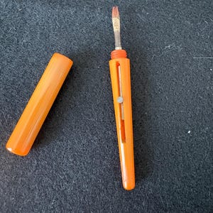 Könnte beinhalten: Ein leuchtend orangefarbener, einziehbarer Pinsel mit passender abnehmbarer Kappe. Der Griff verfügt über einen vertikalen Schlitz mit einem kleinen Metallschieber zum Aus- oder Einfahren des Pinselkopfes, der hellbraune Borsten und eine silberne Metallzwinge hat.