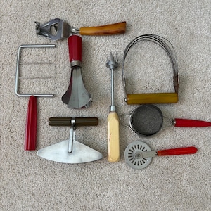 May include: A collection of vintage kitchen utensils including a wire cheese slicer with a red handle, a metal pastry blender with a yellow handle, a red-handled egg slicer, a small mesh strainer, a pizza cutter, and various metal food tools.