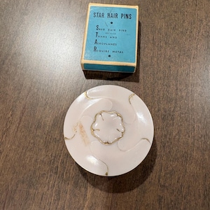 Puede incluir: Una caja vintage de "STAR HAIR PINS" con texto sobre fondo azul, junto a un compacto redondo y blanco con un diseño floral dorado. El compacto tiene un borde dorado y un tono rosado.