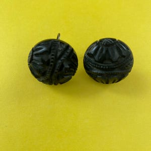 May include: Two black, intricately carved round beads with detailed patterns. One bead has a small metal loop attached. The beads are set against a bright yellow background, highlighting their dark color and textured surfaces.