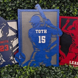 May include: Three wooden sports-themed wall art pieces. One features a basketball player with the name "SMITH" and the number "23". Another depicts a baseball player with "TOTH" and "15". The third shows a volleyball player with "LEAH" and the number "1".