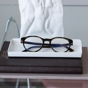 May include: A white rectangular tray with a pair of tortoiseshell eyeglasses on top. The tray is sitting on a brown leather-bound journal with the word "aromas" printed on the front.